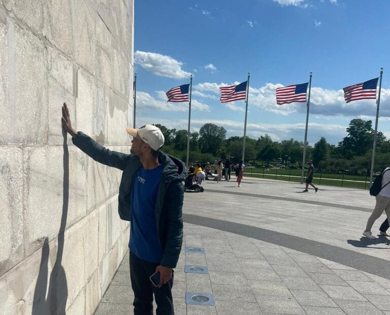 Wil stands with his hand outstretched touching the base of the Washington Monument. Behind him are four American flags.